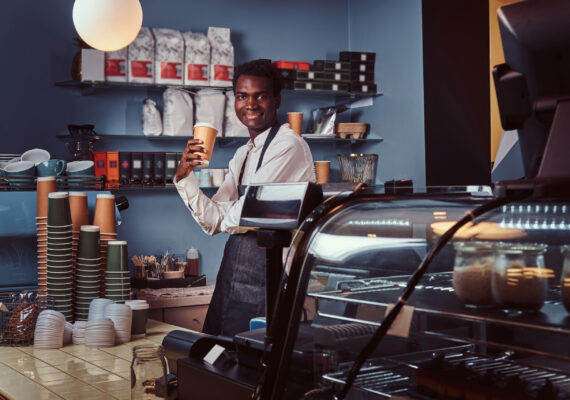 Smiling handsome African American barista in uniform holds cup of coffee while standing in his coffee shop.