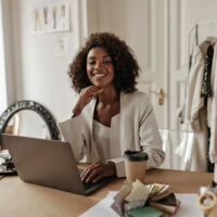 Charming young dark-skinned woman in stylish jacket and blouse smiles, looks at camera, works in laptop and poses in office.